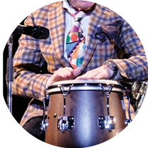 Helge Schneider sitzt im karierten Sakko vor einem Mikrofon und spielt Bongo.