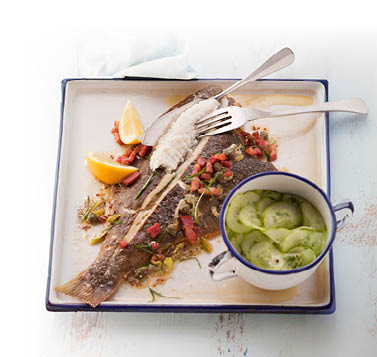 Gebratenes Schollenfilte, angerichtet auf einem quadratischen Teller mit zwei Zitronenspalten, Kapern und Sardellenfilets. Dazu steht eine kleine Tasse Gurkensalat auf dem Teller sowie ein Fischmesser und eine Gabel.