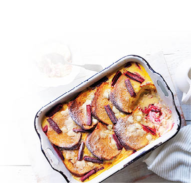 Auf einem rustikalen wei en Holztisch steht eine rechteckige Auflaufform aus Emaille, in der ein s er Auflauf serviert wird. In der Form liegen mehrere goldbraun gebackene Brotscheiben in einer Vanille-Eier-Mischung, durchzogen mit gebackenen St cken von rotem Rhabarber. Der Auflauf ist leicht mit Puderzucker best ubt. Ein St ck wurde bereits entnommen, sodass die weiche, cremige Textur im Inneren sichtbar ist.