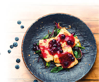 Auf dem Bild sieht man ein liebevoll angerichtetes Essen auf einem schwarzen Teller. Es handelt sich um gebratenen Kabeljau, also wei en Fisch. Der Fisch liegt auf frischem Blattgem se, zum Beispiel Spinat oder Mangold. Dar ber ist eine leuchtend rote Heidelbeerso e gegeben – mit ganzen Beeren darin. Links steht ein Glas mit der So e, daneben liegen frische Heidelbeeren auf dem Holztisch.