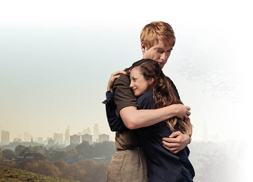 Auf dem Bild umarmen sich ein Mann und eine Frau. Der Mann hat hellrote Haare und tr gt ein graues Hemd. Die Frau hat lange braune Haare und tr gt ein dunkelblaues Oberteil. Sie dr ckt ihr Gesicht an seine Schulter und sieht dabei traurig oder ger hrt aus. Die beiden stehen auf einer Wiese. Im Hintergrund sieht man eine Stadt mit vielen Hochh usern. Es ist hell, der Himmel ist blau.