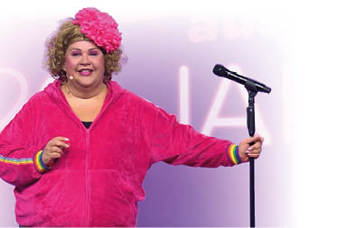 Eine korpulente Frau in einem pinken Jogginganzug h lt sich mit der linken Hand an einem schwarzen Mikrofonst nder fest. Sie hat blonde Locken und eine gro e pinke Blume im Haar stecken.