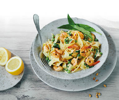Ein tiefer grauer Teller mit einem Pastagericht steht auf einem flachen grauen Teller. Neben breiten langen Nudeln erkennt man gebratene Garnelen und gr ne Bl tter in einer hellen Sauce. Neben dem Pastateller steht ein kleinerer grauer Teller. Darauf liegt die beiden H lften einer aufgeschnittenen Zitrone.