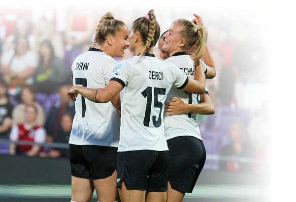 Eine Gruppe von Fu ballspielerinnen liegt sich bei einem Torjubel in den Armen. Die Spielerinnen tragen wei e Trikots und schwarze Hosen. Im Hintergrund sieht man leicht verschwommen Zuschauerreihen.