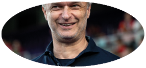 Ein Mann mit grauen Haaren und Drei-Tage-Bart l chelt in die Kamera. Er tr gt ein schwarzes Hemd.