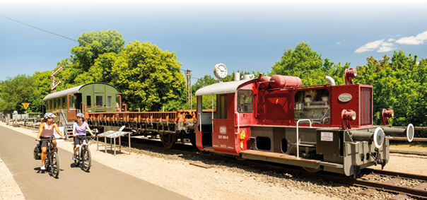 Das Bild zeigt eine sonnige Szene im Freien an einem historischen Bahnhof. Im rechten Bereich des Bildes steht eine kleine rote Diesellok mit der Fahrzeugnummer 323 108-8, die zur Deutschen Bundesbahn geh rt. Der Motorbereich der Lok ist teilweise verglast, sodass technische Bauteile sichtbar sind. Am vorderen Teil der Lok sind wei e Pufferringe und eine Trittstufe montiert. Hinter der Lok ist ein offener, rostbrauner Flachwagen sowie ein gr ner, geschlossener Waggon mit kleinen Fenstern zu sehen. Eine runde Bahnhofsuhr ist auf einem Mast auf dem Wagendach montiert.   Im linken Bildbereich verl uft ein asphaltierter Radweg parallel zu den Bahngleisen. Darauf fahren zwei Frauen nebeneinander auf Fahrr dern. Beide tragen Fahrradhelme und sportliche Kleidung in hellen Farben. Eine der Frauen hat Fahrradtaschen am Gep cktr ger befestigt. Sie fahren entspannt nebeneinander und unterhalten sich offenbar.    Der Hintergrund ist gepr gt von  ppigem, gr nem Laub und kr ftig belaubten B umen. Der Himmel ist strahlend blau mit nur wenigen kleinen wei en Wolken. Die Szenerie wirkt sommerlich und freundlich. Am linken Bildrand stehen mehrere historische Eisenbahnsignale und Informationstafeln neben dem Gleis. Der gesamte Eindruck des Bildes ist hell, klar und ruhig – es vermittelt eine harmonische Verbindung von Natur, Technikgeschichte und Freizeitaktivit t.
