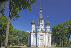 Das Bild zeigt eine russisch-orthodoxe Kirche mit blauer Holzfassade und wei en Balken. Die Kirche hat 3 T rme, welche jeweils ei goldenen Kreuz an der Spitze haben. Die Kirche ist umgeben von B umen, der Himmel ist klar blau. 