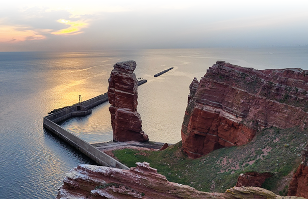 Das Bild zeigt eine beeindruckende Felsformation an der K ste bei Sonnenuntergang. Im Vordergrund und rechts sind rote, schichtweise aufgebaute Klippen zu sehen, die von etwas gr ner Vegetation bedeckt sind. Zentral im Bild ragt ein hoher, schlanker Felsen steil aus dem Wasser empor, umgeben von einem geschwungenen Schutzdamm oder Hafenmole, die das Wasser teilweise einf ngt und abgrenzt. Der Himmel ist teils bew lkt, wobei die Sonne hinter den Wolken teilweise sichtbar ist und das Meer mit einem goldenen Schimmer berzieht. Die Stimmung wirkt ruhig und friedlich, die Farben warm und harmonisch. Im Hintergrund ist das weite Meer zu sehen, das bis zum Horizont reicht.  