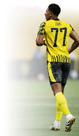 Ein junger Fu ballspieler ist auf dem Spielfeld zu sehen. Er tr gt ein gelb-schwarzes Trikot mit der R ckennummer 77 und dem Namen “Jobe" auf dem R cken. Die Trikot rmel und die R ckseite des Trikots haben ein auff lliges, schwarzes Muster. Die Shorts sind schwarz mit grauen Mustern, und an den Seiten verlaufen gelbe Streifen. Der Spieler tr gt gelbe Stutzen und wei e Fu ballschuhe mit schwarzen Streifen und goldener Sohle. Er befindet sich in einer laufenden Bewegung auf einem gr nen Fu ballfeld, der Hintergrund ist unscharf, aber es sind Zuschauer und eine gelbe Stadionwand zu erkennen.