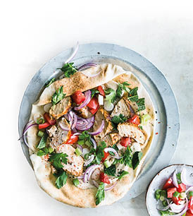 Auf einem runden Metallteller liegt ein aufgeschlagenes, weiches Fladenbrot. Darauf sind St cke von gegrilltem H hnchenfleisch, frische Tomatenw rfel, rote Zwiebelringe, geschnittene Fr hlingszwiebeln und gr ne Kr uter wie Petersilie und Minze verteilt. Unter den Zutaten ist eine cremige, helle Sauce zu sehen. Links oben im Bild steht eine kleine Schale mit einer weiteren Portion der Sauce, daneben ein kleiner Teller mit zus tzlichem Tomaten-Zwiebel-Salat. Die Szene wirkt frisch, farbenfroh und appetitlich.