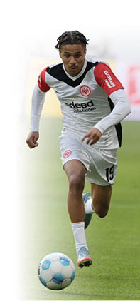 Ein junger m nnlicher Fu ballspieler in Aktion auf dem Spielfeld. Er tr gt ein wei es Trikot mit roten und schwarzen Akzenten, das Logo von Eintracht Frankfurt ist auf der Brust zu sehen. Auf dem linken rmel steht der Schriftzug „ELOTRANS Reload“. Die Trikotnummer „19“ ist auf seiner linken Hose zu erkennen. Der Spieler hat kurze, geflochtene Z pfe und konzentriert sich auf den Ball vor ihm, den er offenbar dribbeln will. Er tr gt wei e Stutzen und hellblaue Fu ballschuhe mit pinken Akzenten. Im Hintergrund ist ein weiterer Spieler in einem dunkleren Trikot zu sehen, aber unscharf. Die Szene findet auf einem gr nen Rasenplatz statt.