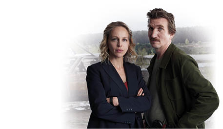 Im Vordergrund stehen eine Frau und ein Mann nebeneinander. Die Frau tr gt einen dunklen Blazer, hat blonde, offene Haare und blickt ernst in die Kamera. Der Mann tr gt eine gr ne Jacke, hat braune Haare und einen Schnurrbart, auch er schaut ernst. Im Hintergrund wird ein stark verbranntes Auto mit einem Kran aus dem Wasser gezogen. Mehrere Personen in wei en Schutzanz gen sowie ein Polizeifahrzeug sind am Einsatzort zu sehen. Die Umgebung wirkt l ndlich, mit Wald und Wasserfl che.