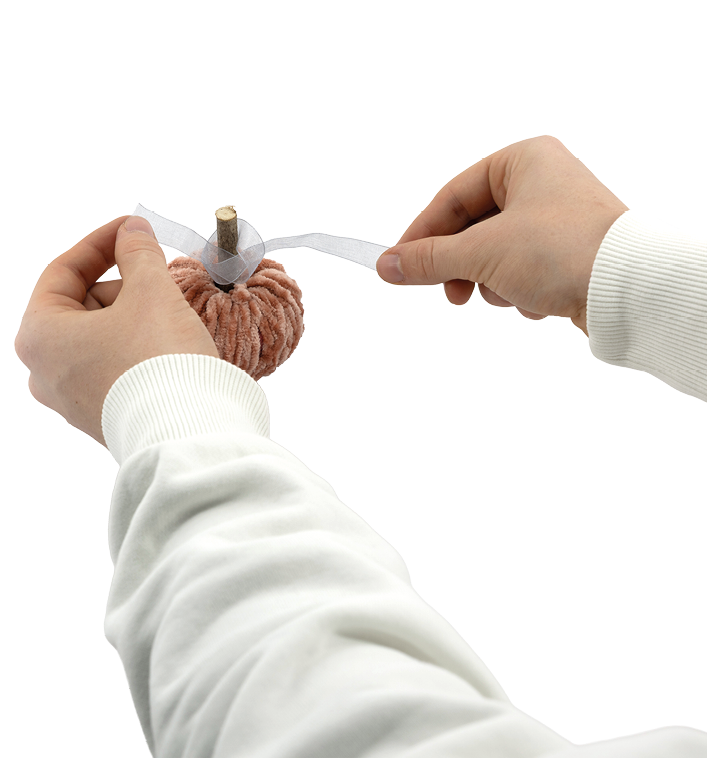 Auf dem Bild ist zu sehen, wie zwei H nde ein kleines, handgefertigtes Objekt verzieren, das wie ein K rbis aussieht. Der K rbis besteht aus flauschiger, orangefarbener Wolle, und oben steckt ein kurzer Holzstab als Stiel. Die Person bindet gerade ein durchsichtiges, helles Band (vermutlich Organza oder ein hnliches Material) um den Stiel, um dem K rbis eine dekorative Schleife zu verleihen. 