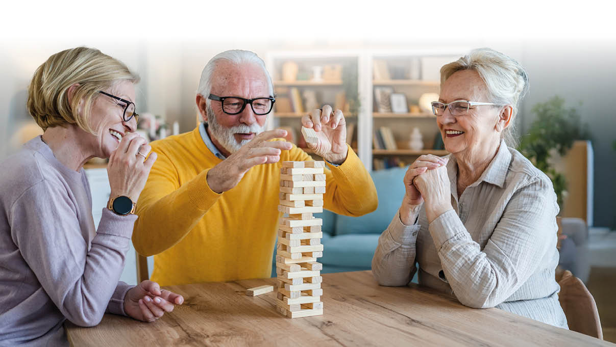 Das Bild zeigt drei ltere Menschen, die gemeinsam an einem Tisch sitzen und Jenga spielen. Alle drei wirken fr hlich und konzentriert – es wird gelacht, gestaunt und mitgefiebert. Die Szene spielt offenbar in einem hellen, gem tlichen Wohnzimmer mit Pflanzen und B cherregalen im Hintergrund.    Die Atmosph re strahlt Lebensfreude, Geselligkeit und geistige Aktivit t aus – perfekt geeignet f r Themen rund um aktive Senioren, Gemeinschaft, Freizeitgestaltung oder Gehirntraining im Alter.    