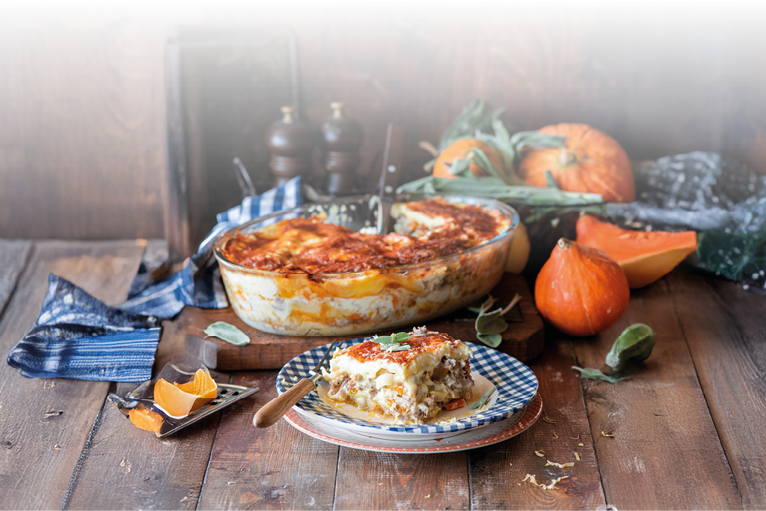 Das Bild zeigt eine herbstlich angerichtete K rbis-Lasagne – k stlich berbacken mit goldbrauner K sekruste. Im Vordergrund liegt ein angerichtetes St ck auf einem Teller mit blau-wei em Muster, daneben ein rustikales Holzmesser. Im Hintergrund stehen K rbisse, frische Salbeibl tter und eine Auflaufform, aus der bereits Portionen entnommen wurden.