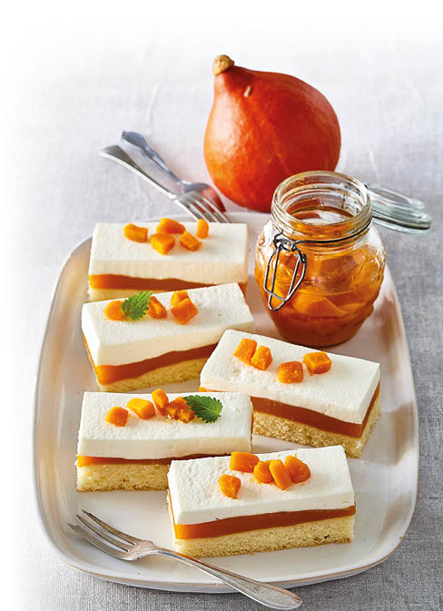 Das Bild zeigt ein herbstliches Dessert – K rbis-Schnitten mit Creme-Schicht. Auf einer rechteckigen Platte liegen mehrere St cke eines feinen Kuchens, bestehend aus drei Schichten:      einem hellen Biskuit- oder R hrteigboden,      einer leuchtend orangefarbenen K rbiscreme,      und einer dicken Schicht heller Sahne- oder Frischk secreme.      Obenauf sind kleine W rfel aus kandiertem oder eingekochtem K rbis sowie etwas Zitronenmelisse als Garnitur verteilt.  Daneben steht ein Glas mit K rbisst ckchen in Sirup – vermutlich selbst eingekocht.  Im Hintergrund liegt ein ganzer Hokkaidok rbis, der farblich perfekt zu den Dessertst cken passt.  Die Komposition wirkt appetitlich, saisonal und elegant angerichtet – ideal f r ein herbstliches Kaffeetrinken oder ein feines Buffet.