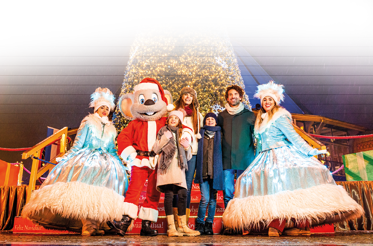 Das Bild zeigt eine fr hliche Winterszene vor einem sehr gro en, reich beleuchteten Weihnachtsbaum. In der Mitte steht eine gro e Maus-Maskottchenfigur im Weihnachtsmannkost m. Links und rechts davon posieren zwei Darstellerinnen in funkelnden, silbrig-blauen, weit ausgestellten Winterkleidern mit wei en Fellbes tzen und gro en, flauschigen Kopfbedeckungen.   Vor und neben dem Maskottchen steht eine Familie: zwei Erwachsene und zwei Kinder, warm in Winterm ntel, M tzen und Schals gekleidet. Alle l cheln und wirken begeistert. Im Hintergrund leuchten bunte Geschenkpakete und festliche Dekorationen, und es scheint leicht zu schneien. Der Boden ist nass und reflektiert die hellen Lichter der Umgebung.