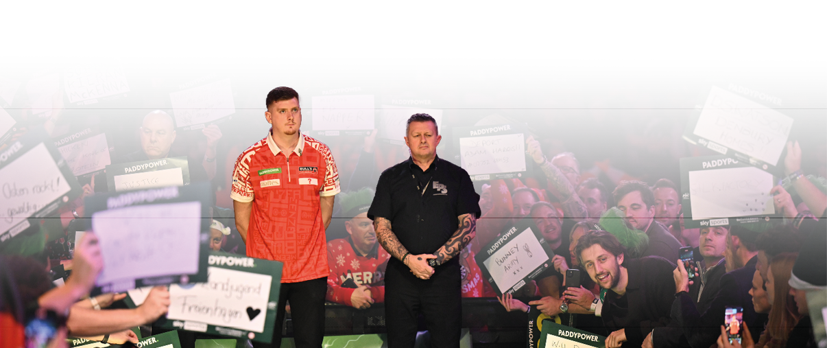 Im ber hmten “Ally Pally" stehen zwei Dartspieler auf der B hne. Der Spieler links tr gt ein rotes Shirt, der Spieler rechts ein schwarzes Shirt. Um die B hne herum stehen Fans, die Schilder hochhalten oder mit dem Handy Fotos machen.