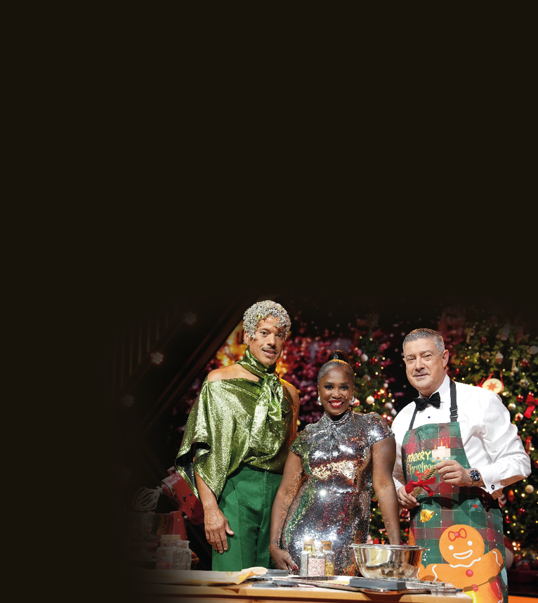 Auf dem Bild stehen drei Personen in festlicher Studio-Kulisse vor weihnachtlich geschm ckten Tannenb umen und berufenen Lichtern. Alle drei sind elegant bzw. auff llig gekleidet: • Der Mann links tr gt ein gl nzendes, gr nes Oberteil mit Schal-Element und eine gr ne Hose; sein Kopf ist mit silbrigem Glitzerschmuck dekoriert.  • Die Frau in der Mitte tr gt ein figurbetontes Kleid aus silbernen, stark reflektierenden Pailletten.  • Der Mann rechts tr gt Hemd, Fliege und eine weihnachtliche Sch rze mit „Merry Christmas“-Aufdruck.  Vor ihnen befindet sich eine Arbeitsfl che mit Back- und Kochutensilien wie Sch sseln, Gew rzgl sern und einem Mixer.   