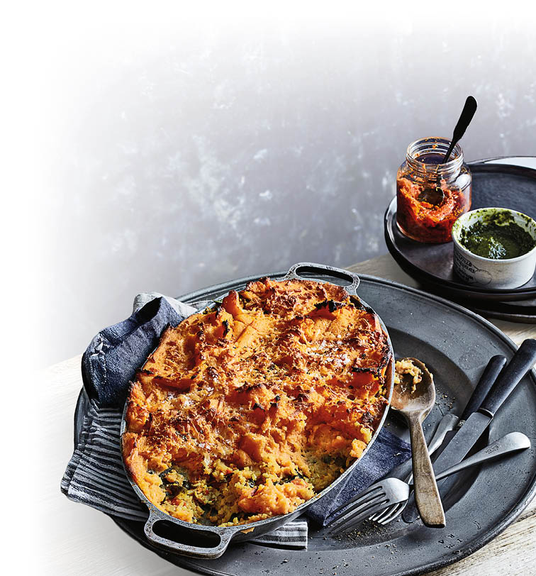 Auf einem gro en Metalltablett steht eine Auflaufform mit einem goldbraun gebackenen Auflauf, daneben liegen ein L ffel und eine Gabel, im Hintergrund stehen ein Glas mit roter Sauce und eine Schale mit gr ner Sauce.