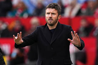 Mann in schwarzem Mantel mit erhobenen H nden vor unscharfem Hintergrund mit Menschen und rotem Banner