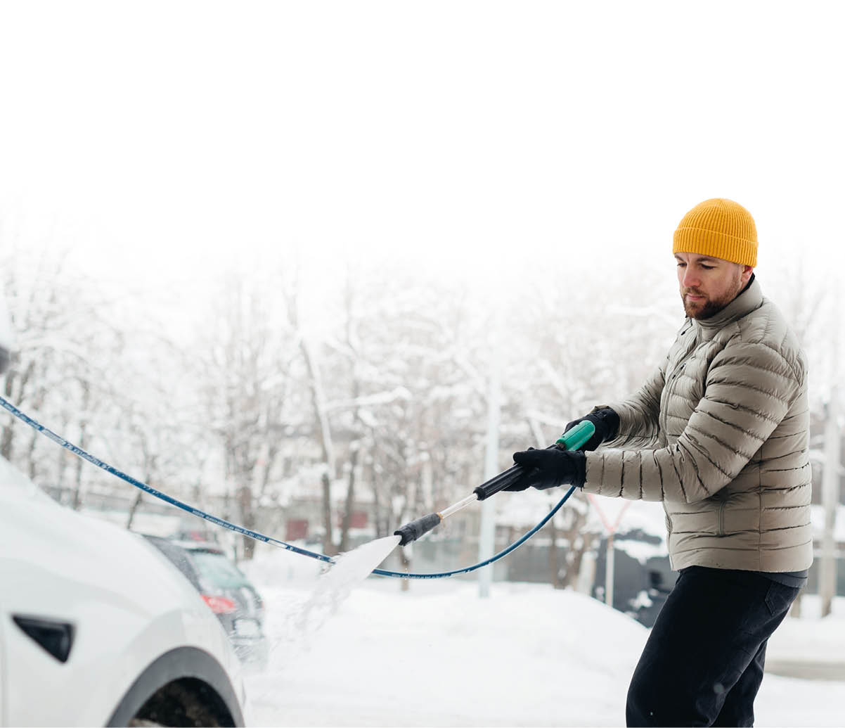 Man cleaning a white electric vehicle with a high-pressure washer during winter, showing car maintenance in cold weather