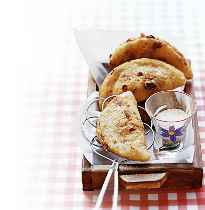 Auf einem rot-wei karierten Tischtuch steht ein kleines rechteckiges Holztablett. Darauf liegen mehrere halbmondf rmige, goldbraun gebackene Teigtaschen, die mit Zucker bestreut sind. Einige der Teigtaschen sind in einer Metallzange oder einem Drahtgestell gehalten. Neben den Teigtaschen steht ein kleines Glas, das mit wei em Zucker gef llt ist und mit einer blauen Blume verziert ist. Das Tablett ist mit wei em Papier ausgelegt. Der Hintergrund ist unscharf, der Fokus liegt auf den Backwaren im Vordergrund.
