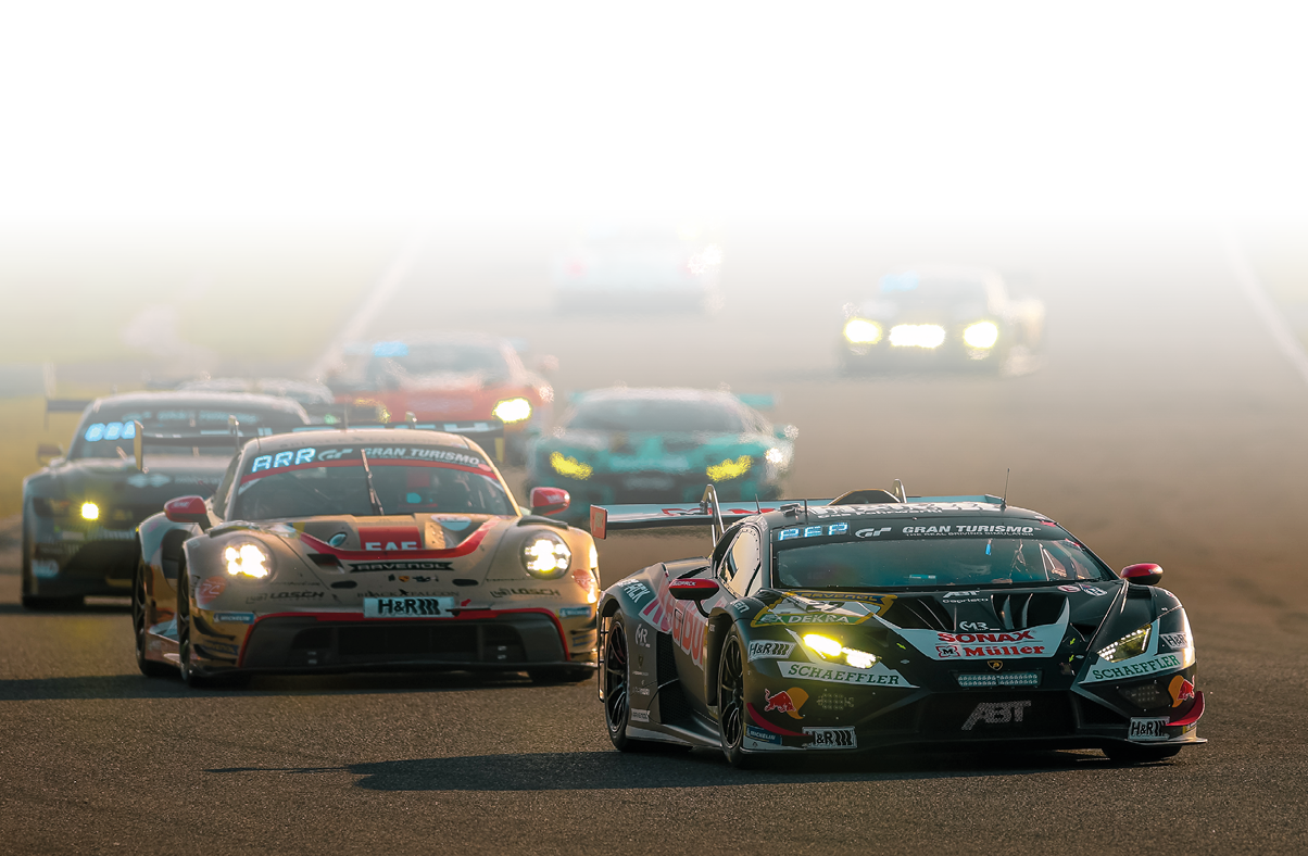 Im Vordergrund fahren mehrere flache, breite Rennwagen (GT-Sportwagen) dicht hintereinander auf einer asphaltierten Strecke. Das f hrende Fahrzeug ist schwarz mit gr nen und roten Sponsorenaufklebern und hat eingeschaltete Scheinwerfer. Direkt dahinter folgt ein heller Rennwagen mit roten Akzenten. Weitere Fahrzeuge sind im Hintergrund zu sehen, teilweise unscharf, was die hohe Geschwindigkeit und Bewegung unterstreicht.    Das Licht ist warm, vermutlich sp ter Nachmittag oder Abend, wodurch die Szene in goldene Farbt ne getaucht ist.      