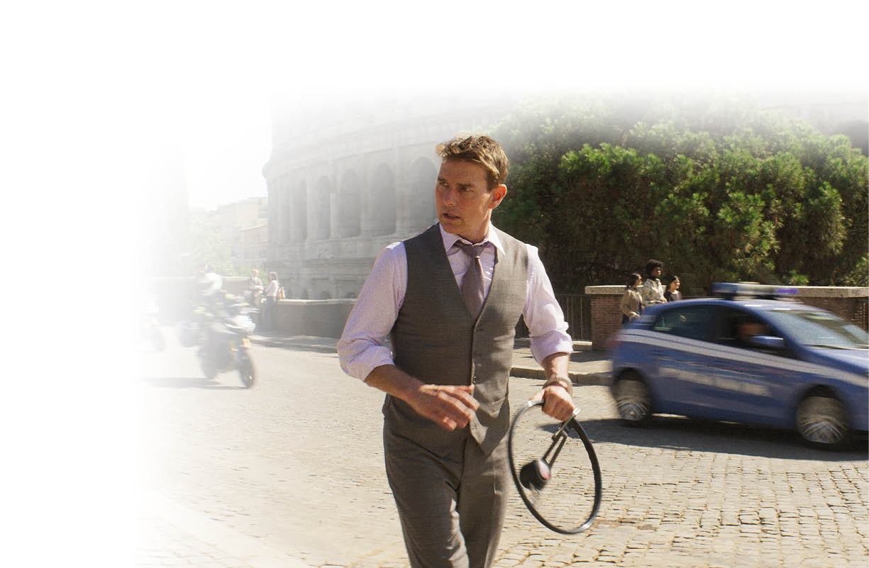 Das Bild zeigt einen Mann in Bewegung auf einer Stra e in einer historischen Stadt.   Er tr gt ein helles Hemd, eine Weste und eine Krawatte und wirkt elegant gekleidet. In einer Hand h lt er Kopfh rer, w hrend er sich z gig nach vorne bewegt. Sein Blick ist nach rechts gerichtet, als w rde er auf etwas reagieren oder sich orientieren.    Im Hintergrund ist deutlich das Kolosseum in Rom zu erkennen. Die Umgebung zeigt eine st dtische Szene mit Kopfsteinpflaster, vorbeifahrenden Fahrzeugen und einigen Menschen am Stra enrand.    Rechts f hrt ein blaues Polizeiauto vorbei, dessen Bewegung leicht verschwommen ist, was Dynamik vermittelt. Auch ein Motorrad ist im Hintergrund zu sehen.    Die linke Bildseite ist stark aufgehellt bzw. ausgeblendet, wodurch der Fokus auf dem Mann und der Szene rechts liegt.