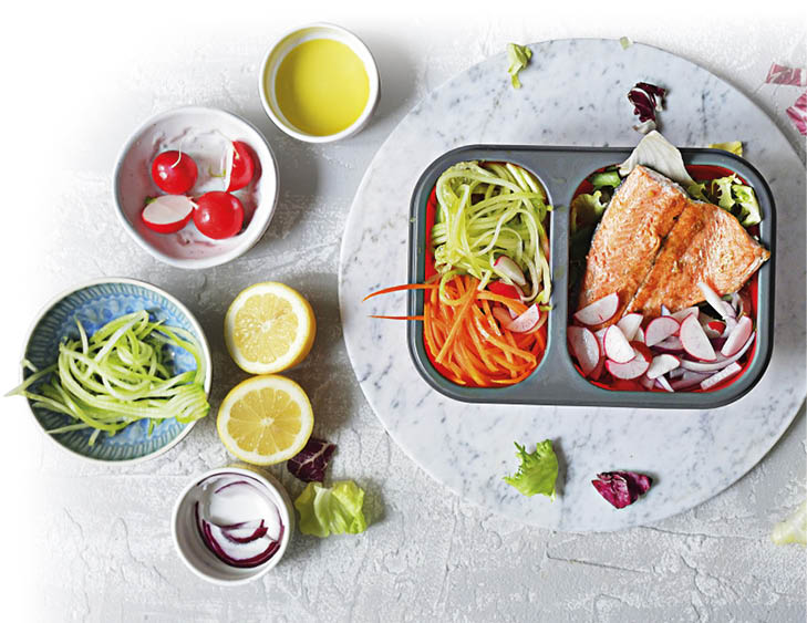 Das Bild ist eine Draufsicht auf eine vorbereitete Mahlzeit in einer rechteckigen Bento‑Box auf einem runden, wei en Marmorteller. In der rechten Box‑H lfte liegt ein gebratenes St ck Lachs auf Salat, daneben sind viele in Scheiben geschnittene Radieschen. In der linken H lfte liegen Gem se‑Streifen: hellgr ne Zucchini oder Gurke und orange M hren, gemischt mit einigen Radieschenscheiben. Rundherum stehen kleine Sch lchen mit Zutaten, darunter ganze Radieschen, Zwiebelstreifen und weitere Gem sestreifen, sowie zwei halbe Zitronen. Oben links steht ein kleines Sch lchen mit gelbem l oder Dressing; einzelne Salatbl tter liegen locker auf dem hellen Tisch verstreut. Personen und Text sind nicht zu sehen.