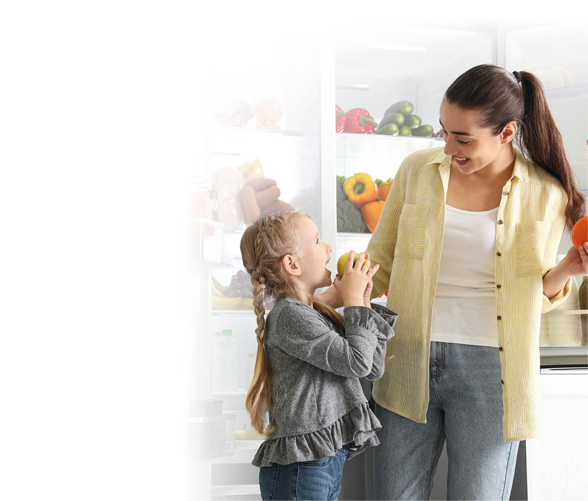 Young mother and her daughter with fruits near open refrigerator in kitchen