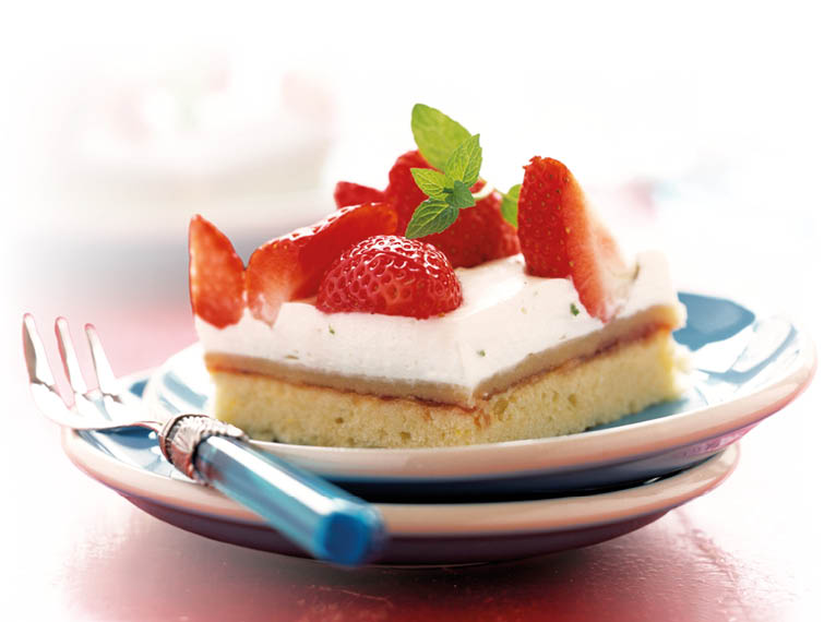 Das Bild zeigt ein St ck Erdbeerkuchen auf einem kleinen Teller. Der Kuchen besteht aus einem hellen, luftigen Boden, darauf eine cremige, wei e Schicht und oben mehrere frische, rote Erdbeeren. Die Erdbeeren sind teils halbiert und gl nzen leicht. In der Mitte liegt ein kleiner gr ner Minzzweig als Dekoration.  Der Teller steht auf einem roten Tisch. Daneben liegen eine Gabel und ein Messer mit blauem Griff.  Im Hintergrund ist ein weiterer Teller mit einem zweiten St ck Kuchen zu sehen, jedoch unscharf.  Die Lichtstimmung ist hell und freundlich, wodurch die Farben besonders frisch wirken.  Gesamteindruck:  Das Bild wirkt appetitlich und sommerlich – ein frisches Dessert mit fruchtiger Note.  