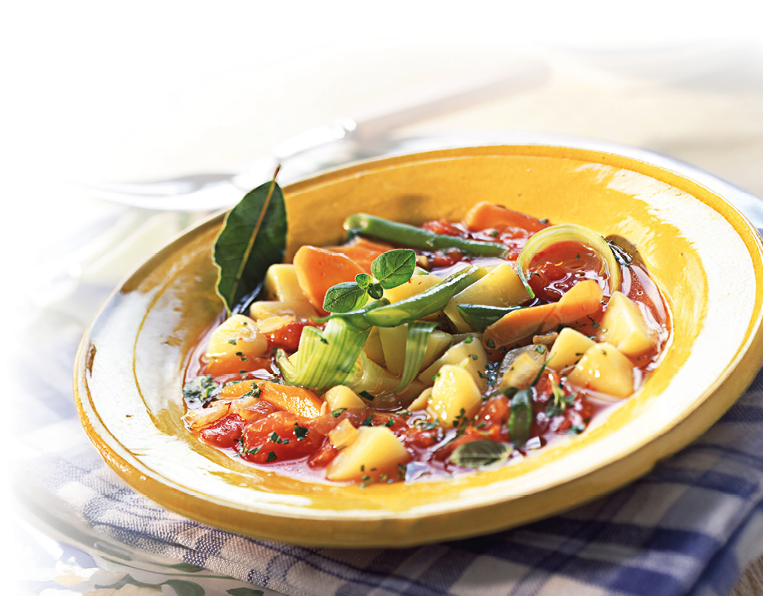 Zu sehen ist eine gelbe, flache Keramikschale mit einer klaren, r tlichen Gem sesuppe. In der Br he liegen gew rfelte Kartoffeln, Karottenscheiben, gr ne Bohnen und Tomatenst cke; dar ber sind frische Kr uter gestreut. Als Garnitur sind ein Lorbeerblatt sowie gr ne Streifen von Lauch oder Fr hlingszwiebel in der Mitte platziert. Die Schale steht auf einem blau-wei karierten Tuch; im hellen Hintergrund sind Teller und Besteck unscharf zu erkennen.