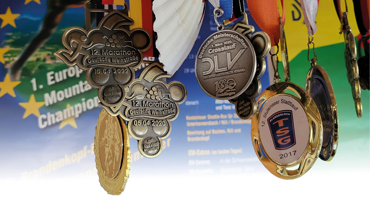 Auf dem Foto h ngen mehrere Laufsport-Medaillen dicht nebeneinander an verschiedenfarbigen B ndern. Die Medaillen sind vorn silbern oder goldfarben und zeigen unterschiedliche Pr gungen: runde Exemplare mit Schrift- und Logo-Bereichen, sowie dekorative Anh nger in Form von stilisierten Weintrauben oder runden Siegeln. Ein Band tr gt die schwarz-rot-goldenen Farben, andere B nder sind blau, orange, rot-wei oder schwarz. Zwischen den Metallmedaillen h ngt mittig eine helle Muschelschale an einem d nnen Band. Einige Medaillen zeigen Jahreszahlen und Veranstaltungsnamen (teilweise nicht vollst ndig lesbar), eine tr gt deutlich sichtbare Gravuren und emblemetypische Elemente wie „Marathon“/„Crosslauf“. Im unscharfen Hintergrund ist ein buntes Plakat mit gelben Sternen auf blauem Feld und weiteren Farbfl chen zu erkennen, das die Medaillen farblich kontrastiert. Die Aufnahme betont Materialtexturen (gl nzendes Metall, gewebte B nder, matte Muschelschale) und vermittelt eine Sammlung von Auszeichnungen aus Laufsport- oder Laufevents.