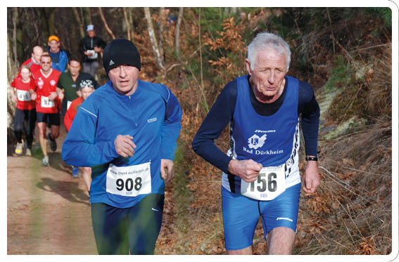 Foto einer Laufszene auf einem naturbelassenen Waldweg. Im Vordergrund laufen zwei Personen nebeneinander: rechts ein lterer Sportler mit grauen Haaren, blaues  rmelloses Trikot  ber einem lang rmeligen schwarzen Laufshirt, knielange blaue Laufshorts und schwarze Laufschuhe; die Startnummer 156 ist an der H fte befestigt. Links daneben ein L ufer in lang rmligem blauem Oberteil und schwarzen Laufleggings, mit wei en Sportschuhen und der Startnummer 908 auf der H fte. Beide sind nach vorn gebeugt in Laufbewegung, die Gesichtspartien sind nicht detailliert erkennbar. Der Weg besteht aus festgetretenem Erd- und Sandboden, entlang des Weges liegen trockenes Laub und vereinzelte Gr ser. Im mittleren und hinteren Bildbereich folgen weitere Teilnehmer in unterschiedlichen Sportoutfits; vereinzelte B ume und Str ucher s umen den Pfad. Das Licht ist hell, vermutlich Tageslicht, und betont die Farben der Laufbekleidung. Die Komposition vermittelt das Tempo und die Anstrengung der L ufer sowie die nat rliche Umgebung des Laufwegs.