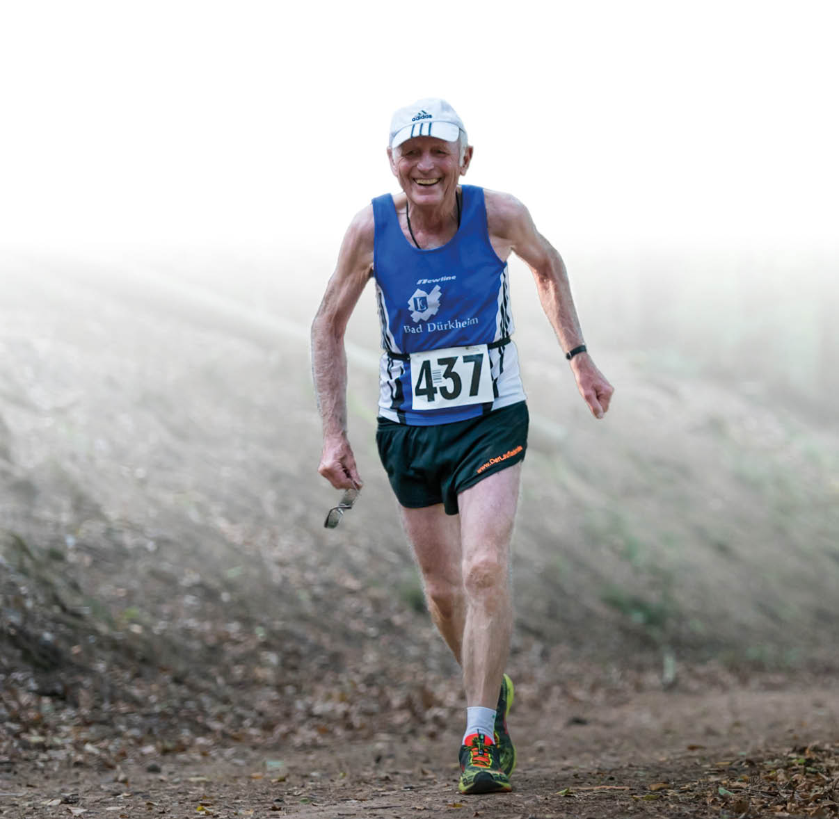 Auf dem Foto ist ein lterer, sportlich wirkender L ufer zu sehen, der auf einem schmalen, leicht abfallenden Waldweg unterwegs ist. Er tr gt ein blaues  rmelloses Lauftrikot mit wei em Logo und der Aufschrift „Bad D rkheim“ sowie die auf der Bauchmitte befestigte Startnummer 437. Dazu hat er kurze schwarze Laufhosen, wei e Sportsocken und bunte Laufschuhe. Auf dem Kopf sitzt eine helle Kappe, an seinem linken Handgelenk ist eine Sportuhr zu erkennen. In der linken Hand h lt er eine dunkle Sonnenbrille. Der Hintergrund zeigt einen bewaldeten Hang mit verschwommenen B umen und weichem, diffusen Licht, der Weg ist mit Laub und kleinen Steinen bedeckt. Die Bildkomposition betont Bewegung nach vorne und die Konzentration des L ufers.