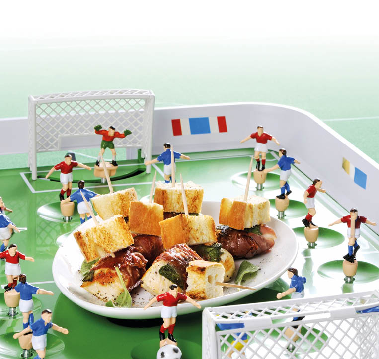 Sehr nah aufgenommene Szene eines Tischfu ballspiels:  In der Bildmitte ein wei er Teller mit einer Portion H hnchenfleischspie en, garniert mit Brotw rfeln und Salbeibl ttern auf Holzspie en. Der Teller steht auf dem gr nen Spielfeld eines Tischfu balls (Kicker). Um den Teller herum stehen kleine Holzfiguren in rot-wei en und blau-wei en Trikots, die typischen Kicker-Spieler, sowie zwei wei e Miniatur-Tore im Vorder- und Hintergrund. Helle, freundliche Farbstimmung.