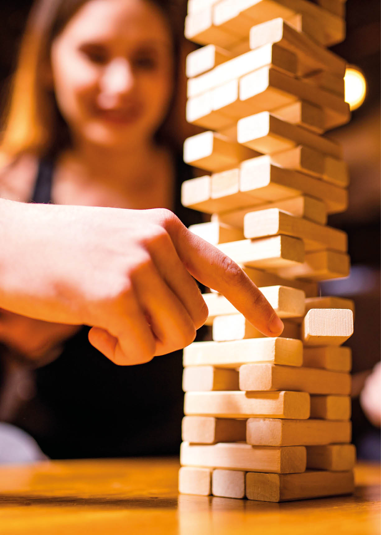 Mehrere Personen spielen in einer gem tlichen Umgebung ein Jenga-Spiel, wobei eine Person gerade einen Holzklotz aus dem Turm zieht. Beschreibung von KI generiert