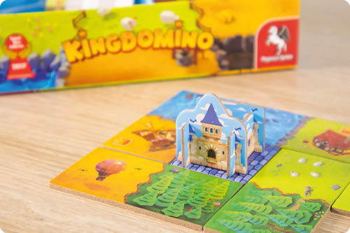 Das Foto zeigt eine Spielsituation des Brettspiels „Kingdomino“ auf einer hellen Holzoberfl che. Im Vordergrund liegen vier quadratische Landschaftspl ttchen zu einem gr eren Quadrat ausgelegt; sie zeigen unterschiedliche Gel ndearten in kr ftigen Farben, darunter gr ne Wald- oder Wiesenfl chen sowie gelbe Felder. In der Mitte steht ein kleines, aufstellbares Papp-Schloss in Hellblau und Beige auf einer angedeuteten Steinfl che. Im Hintergrund ist die Spielschachtel zu sehen, leicht unscharf; darauf ist der Schriftzug „Kingdomino“ in Gelb erkennbar sowie ein rotes Verlagslogo rechts.      