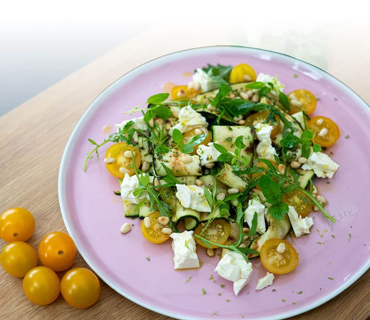 Auf einem rosa Teller liegt ein Salat aus gelben Kirschtomaten, wei en K sew rfeln, gr nen Kr utern und Pinienkernen, daneben liegen mehrere gelbe Kirschtomaten auf einem Holzbrett. Beschreibung von KI generiert