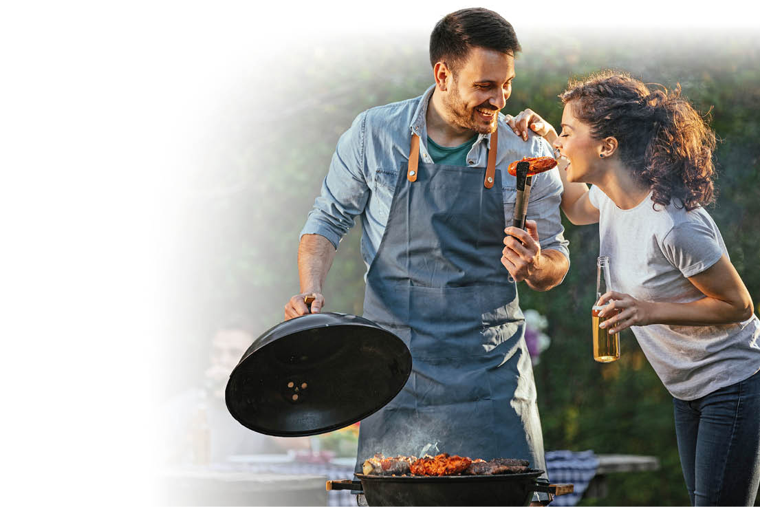 Zwei Personen stehen drau en an einem Grill, eine tr gt eine Sch rze und h lt den Deckel des Grills, w hrend die andere ein Getr nk in der Hand h lt und auf das Grillgut schaut. Beschreibung von KI generiert