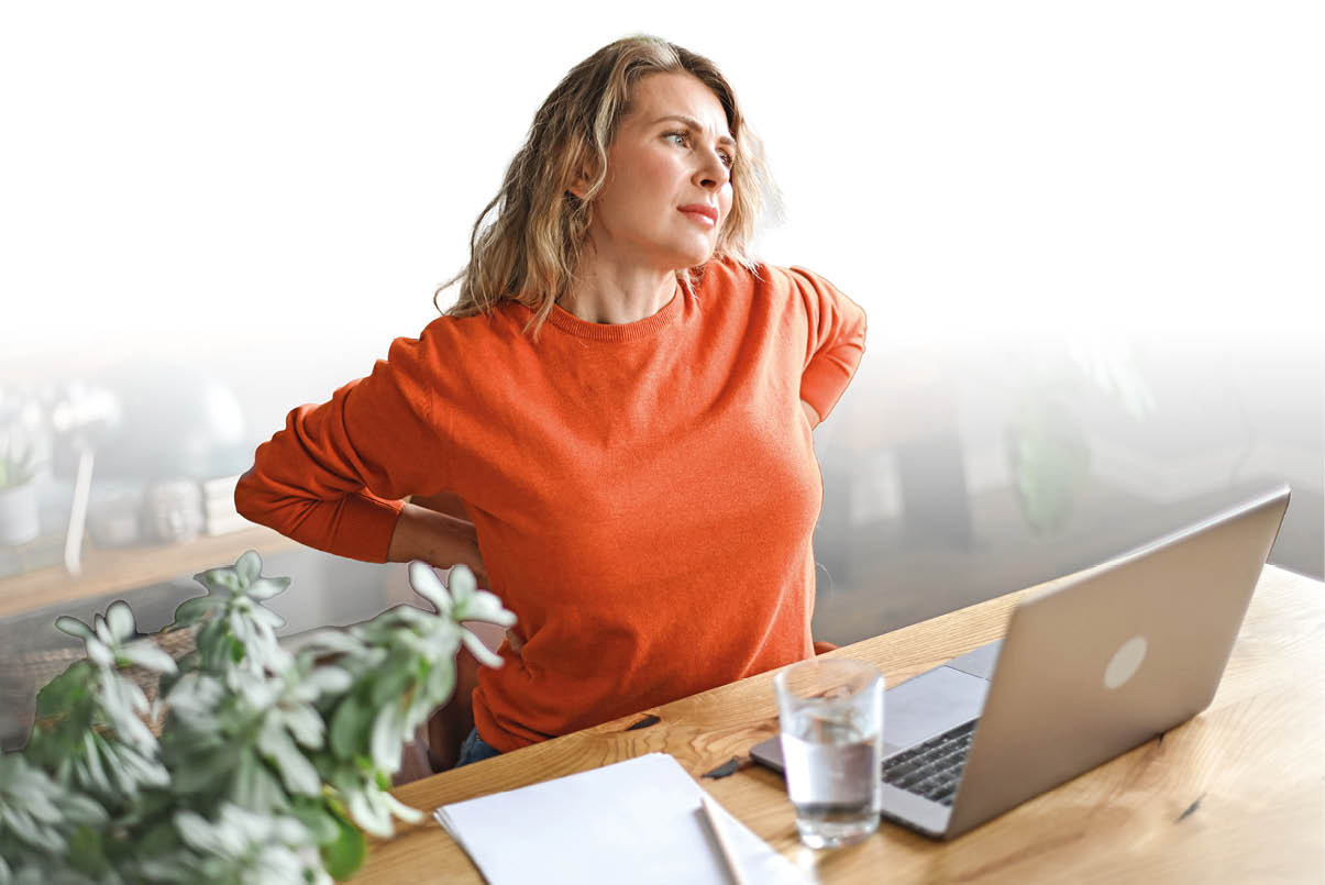 Eine Person mit langen Haaren und einem orangen Pullover sitzt an einem Holztisch vor einem Laptop und h lt sich den R cken, w hrend sie sich zu strecken scheint. Beschreibung von KI generiert