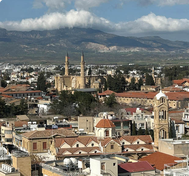 Eine Stadtansicht mit dicht aneinandergebauten Geb uden, mehreren Minaretten und Bergen im Hintergrund unter einem blauen Himmel mit Wolken. Beschreibung von KI generiert