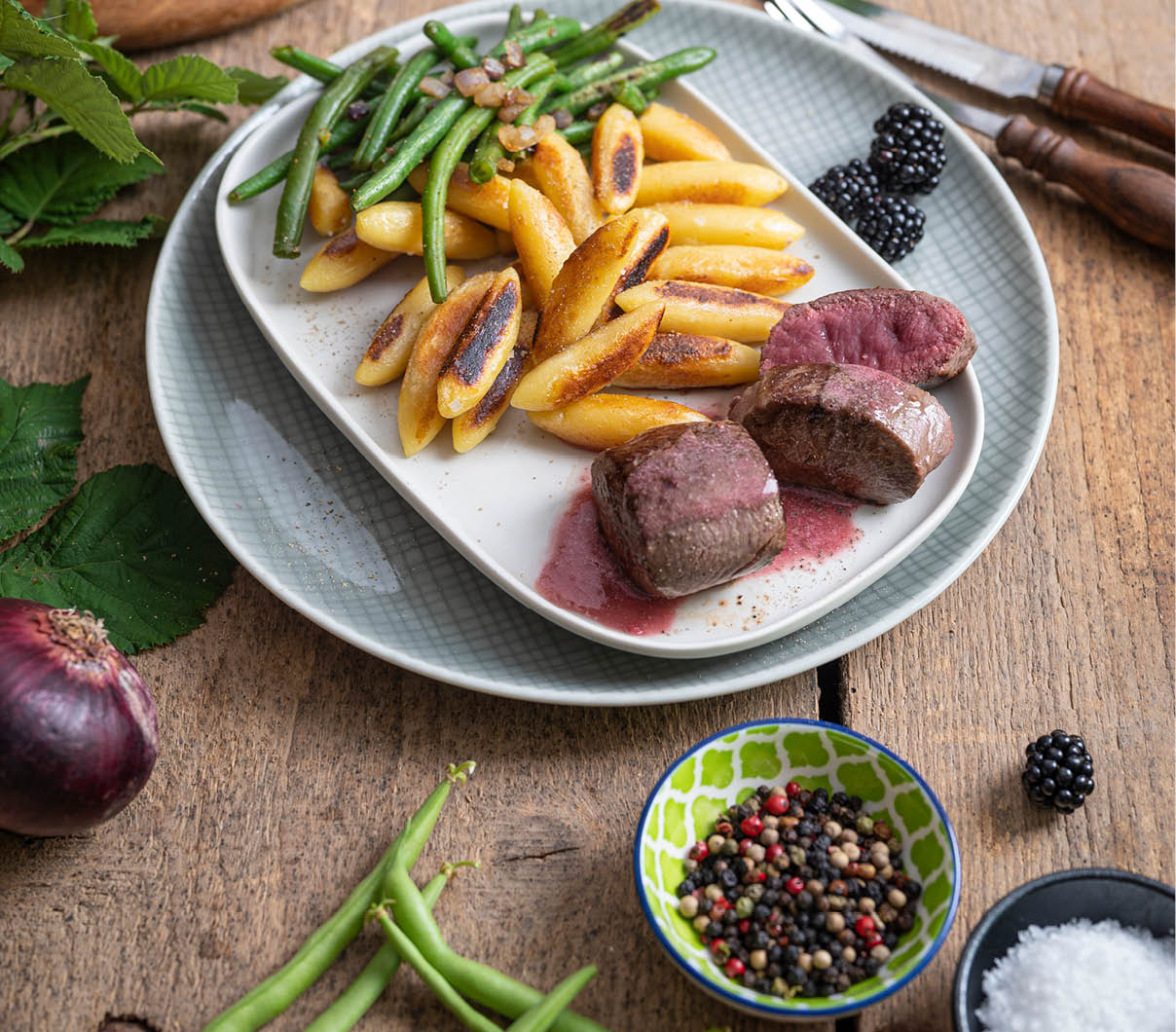 Auf einem Holztisch liegt ein Teller mit gebratenem Fleisch, gr nen Bohnen und gebratenen Kartoffeln, umgeben von frischen Bohnen, Brombeeren, einer Schale mit Pfefferk rnern und einer Schale mit grobem Salz. Beschreibung von KI generiert