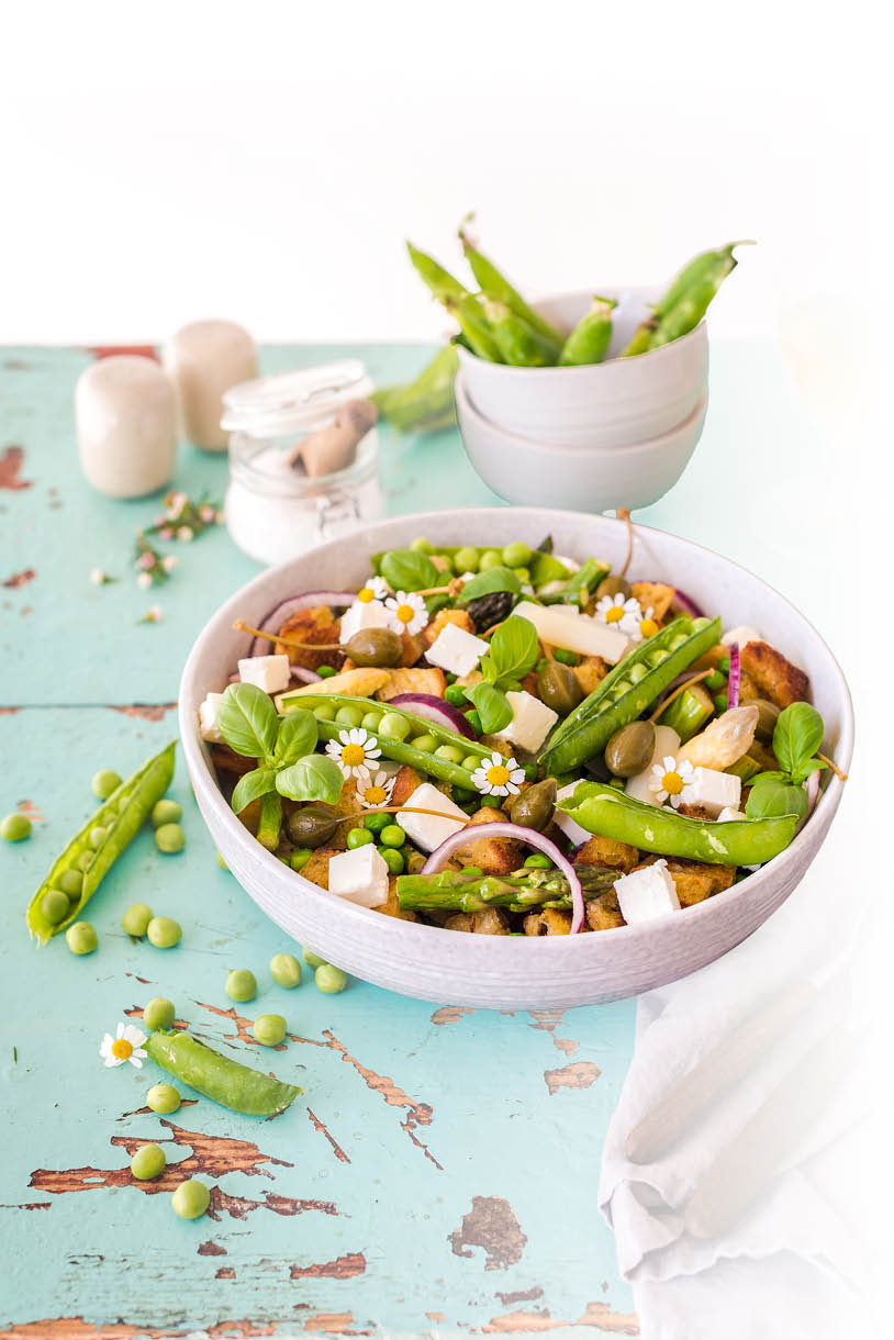 Eine bunte Sch ssel mit frischem Salat aus Erbsen, Feta, Zwiebeln, Kapern und kleinen wei en Bl ten steht auf einem hellblauen, leicht abgenutzten Holztisch neben einer wei en Serviette und weiteren Schalen mit Erbsenschoten. Beschreibung von KI generiert