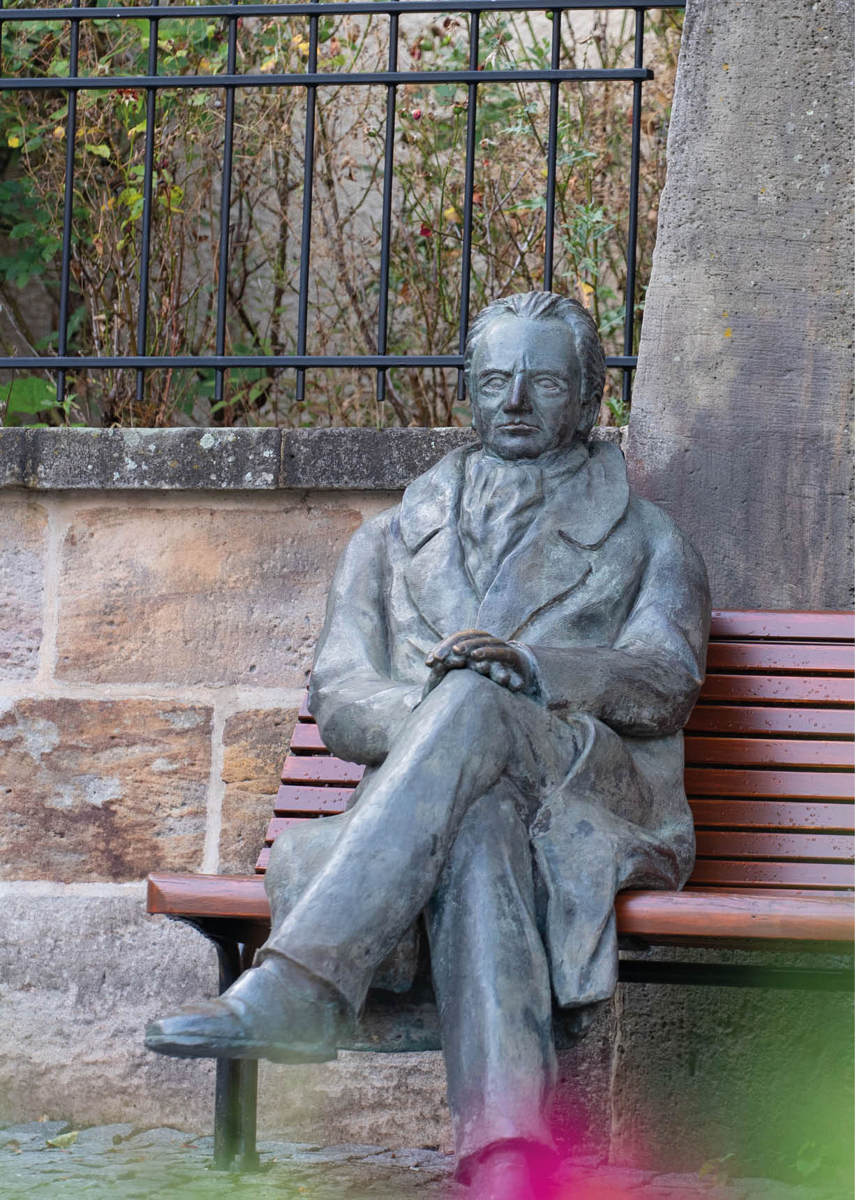Eine Statue einer sitzenden Person mit verschr nkten Beinen und verschr nkten Armen auf einer Parkbank vor einer Steinmauer und einem Metallzaun mit Pflanzen im Hintergrund. Beschreibung von KI generiert