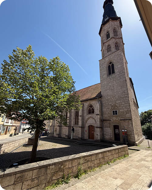 Ein historisches steinernes Geb ude mit einem Turm steht neben einem Baum und einem gepflasterten Weg unter klarem blauem Himmel. Beschreibung von KI generiert