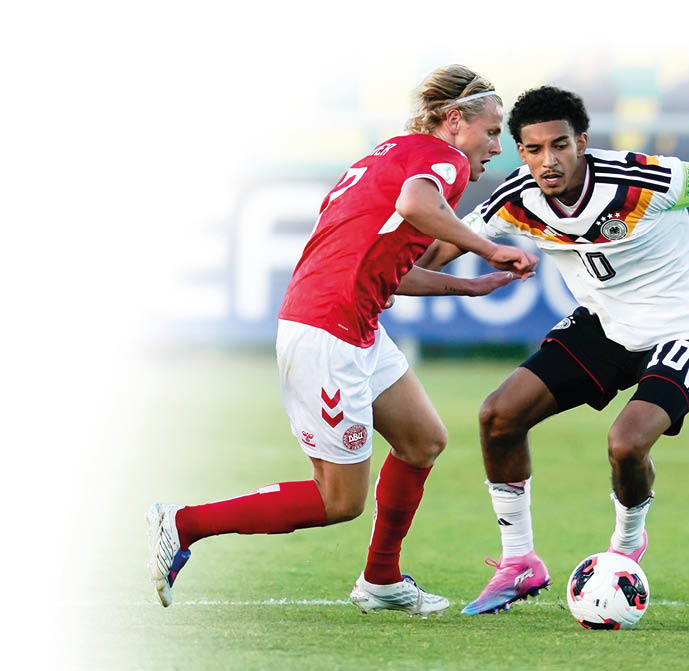 Zwei Erwachsene in Sportkleidung k mpfen auf einem Fu ballfeld um den Ball, wobei einer ein rotes Trikot und der andere ein wei es Trikot mit Kapit nsbinde tr gt. Beschreibung von KI generiert