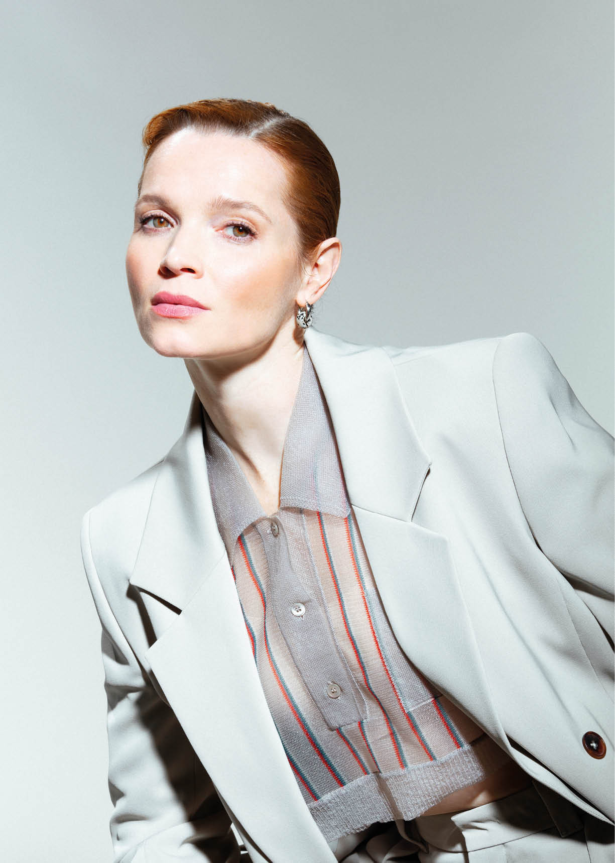 Eine Person mit zur ckgek mmtem Haar tr gt einen hellen Blazer ber einem transparenten, gestreiften Hemd und posiert vor einem hellen Hintergrund.  Beschreibung von KI generiert
