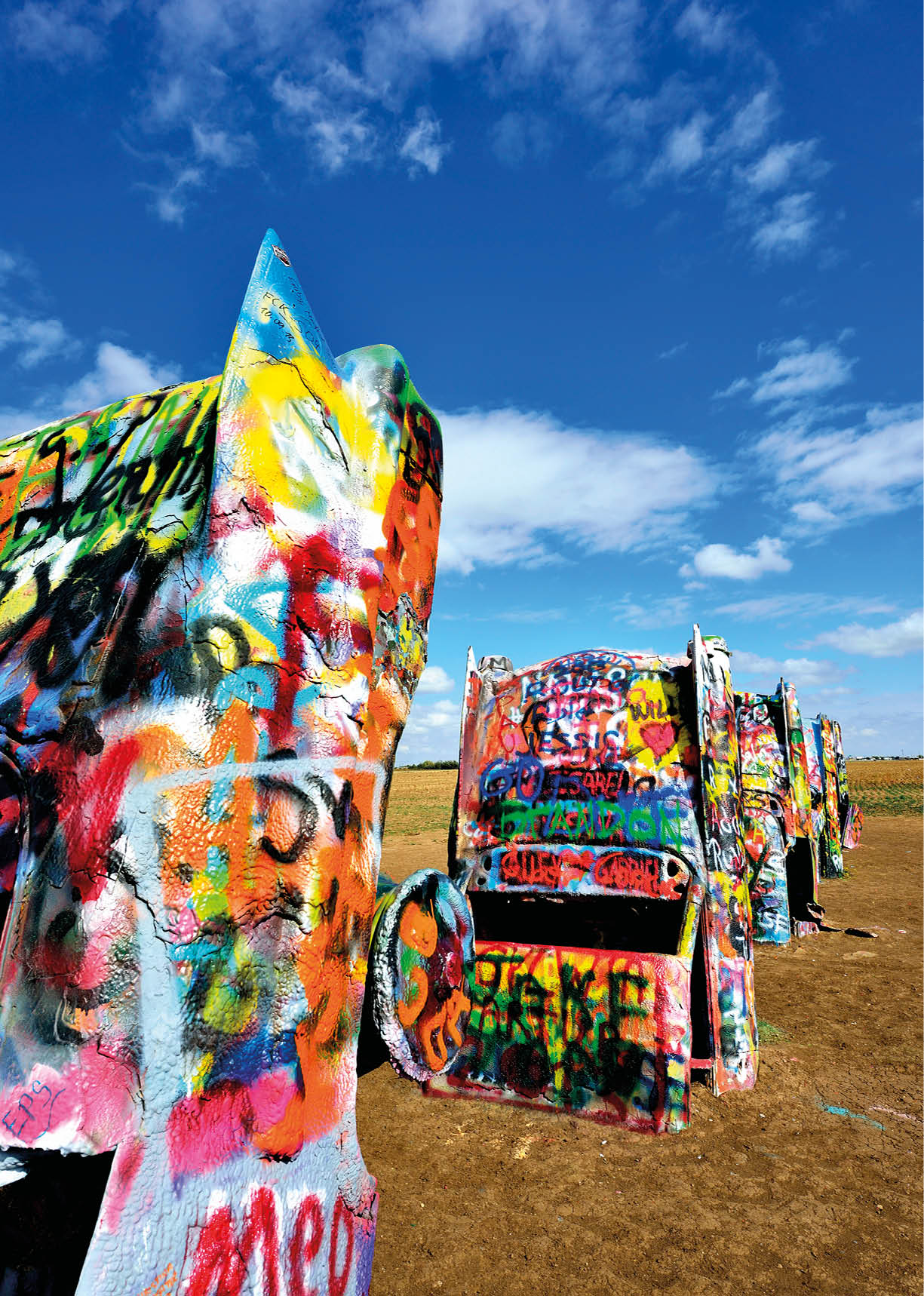 Bunte, mit Graffiti bespr hte alte Autos stehen in einer Reihe auf einem offenen Feld unter einem blauen Himmel mit vereinzelten Wolken. Beschreibung von KI generiert